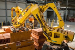 Großes Industriegelände mit gestapelten Kisten, Kränen an der Decke, Zaun, Pfählen und einer fernen Wand in einer automatisierten Fertigungsumgebung.