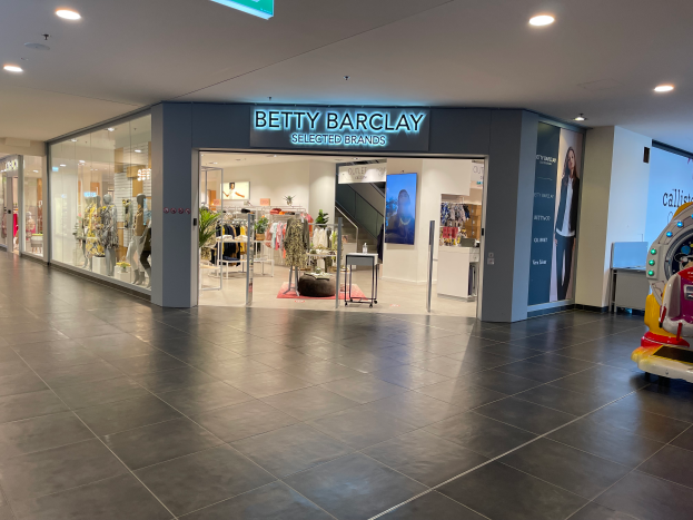 Das Innere eines Betty Barclay-Ladens in einem Einkaufszentrum, mit Schaufensterpuppen in Kleidern, hängenden Kleidungsstücken, Tischen, Pflanzen und anderen Gegenständen, mit einem Spielzeugfahrzeug auf der rechten Seite und einer Tafel mit der Aufschrift "Betty Barclay Selected Brands" oben.