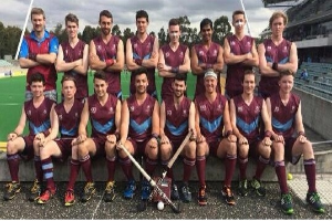 Eine Gruppe von Männern in Sportuniformen steht zusammen auf einem Feld, einige halten Hockeystöcke, feiern mit erhobenen Armen vor einem Stadion mit Zuschauern und einer Kulisse aus Bäumen unter einem klaren blauen Himmel.