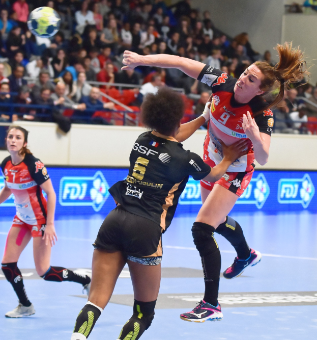 Eine Gruppe von Frauen spielt Handball auf einem Feld, mit einem Ball in der Luft, in einer Stadionatmosphäre, wo Zuschauer auf Stühlen, Geländern und einer Wand sitzen.