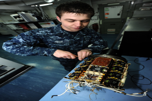 Ein Mann in militärischer Uniform bedient einen Computer an einem unordentlichen Arbeitsplatz mit elektronischen Geräten, umgeben von Maschinen, einem Feuerlöscher, Deckenleuchten und Rohren.