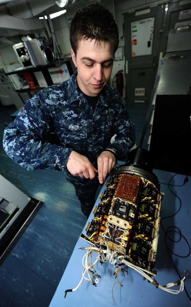 Ein Mann in militärischer Uniform bedient einen Computer an einem unordentlichen Arbeitsplatz mit elektronischen Geräten, umgeben von Maschinen, einem Feuerlöscher, Deckenleuchten und Rohren.