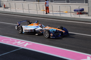 Ein Mann fährt ein Formel-Auto auf einer Rennstrecke, mit einem Helmträger auf dem Fahrersitz, während eine Person auf dem Gehweg im Hintergrund steht, in der Nähe eines Zauns und eines Feuerlöschers.