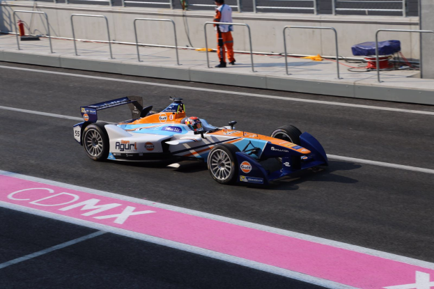 Ein Mann fährt ein Formel-Auto auf einer Rennstrecke, mit einem Helmträger auf dem Fahrersitz, während eine Person auf dem Gehweg im Hintergrund steht, in der Nähe eines Zauns und eines Feuerlöschers.