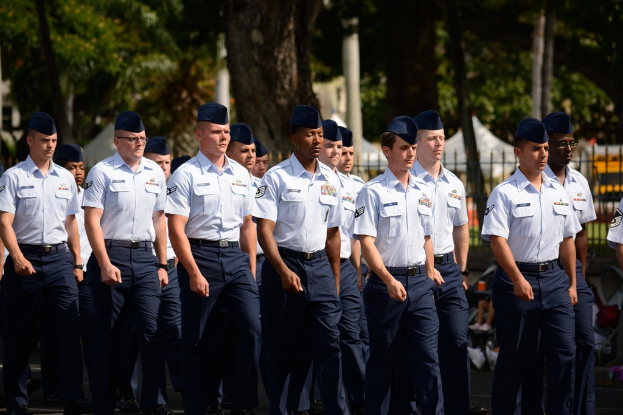 Männer in Uniform gehen auf einer Straße mit einem Eisenzaun dahinter, Bäumen und Polen im Hintergrund.