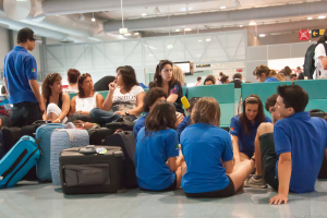 Ein geschäftiger Flughafen-Terminal mit vielen Menschen, die auf ihre Flüge warten, Gepäck auf der linken Seite, Richtungsschilder und Glastüren im Hintergrund.