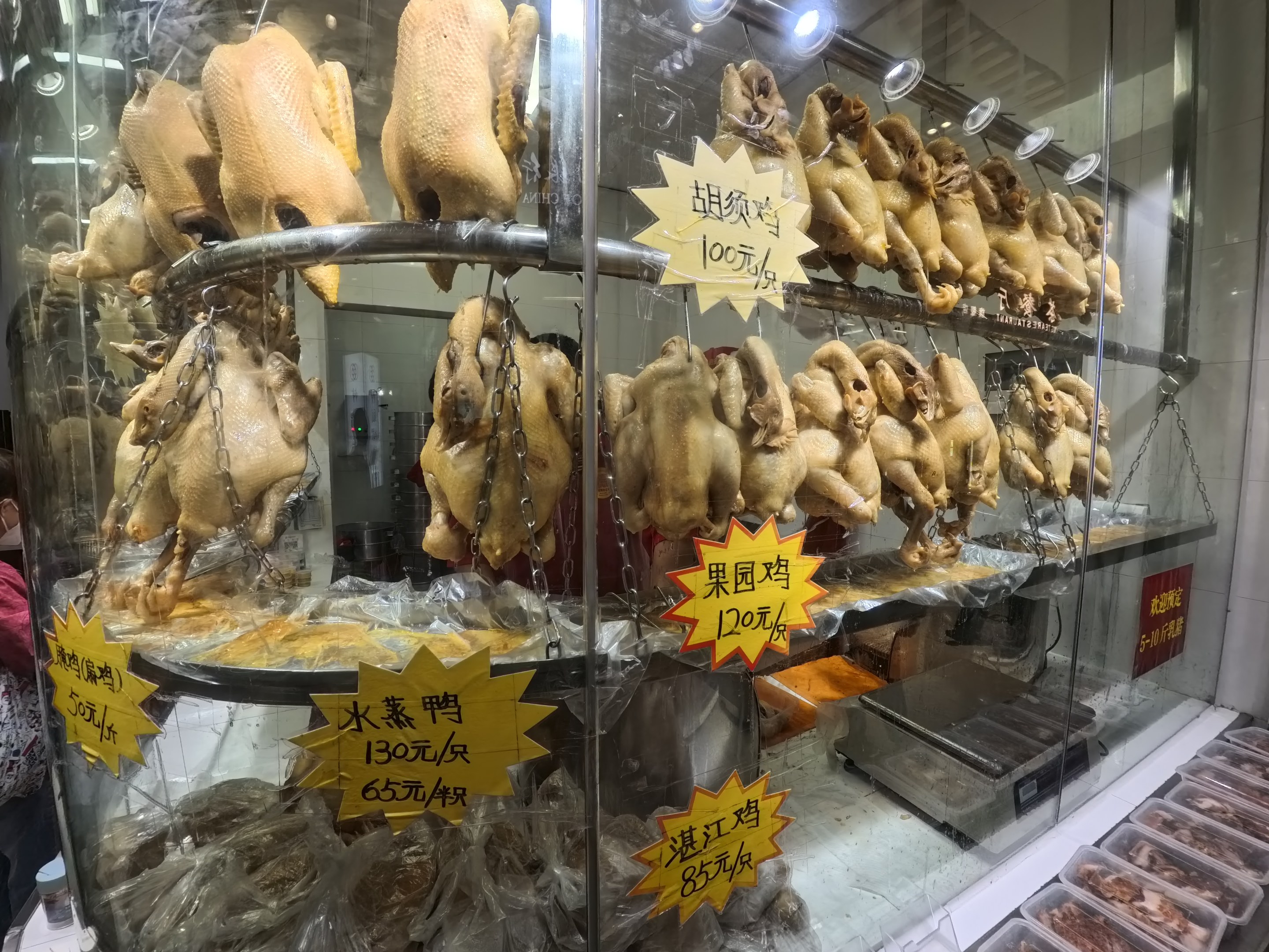 Vitrine mit ganzen gerösteten Hühnern auf einer Theke, mit einer Person links und Tabletts mit Lebensmitteln rechts.