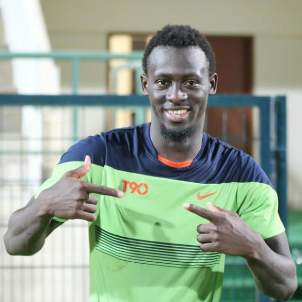 Ein Mann in einem grünen und blauen Hemd, wahrscheinlich Emmanuel Adebayor, zeigt mit einem Lächeln auf die Kamera, mit einem unscharfen Geländer, einer Wand und einer Tür im Hintergrund.