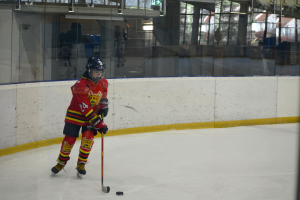 Ein junger Junge mit Helm spielt Hockey auf einem Eisstadion, hält einen Hockeystock und hat Menschen, Bäume, Gebäude und den Himmel durch die umgebende Glaswand im Hintergrund.