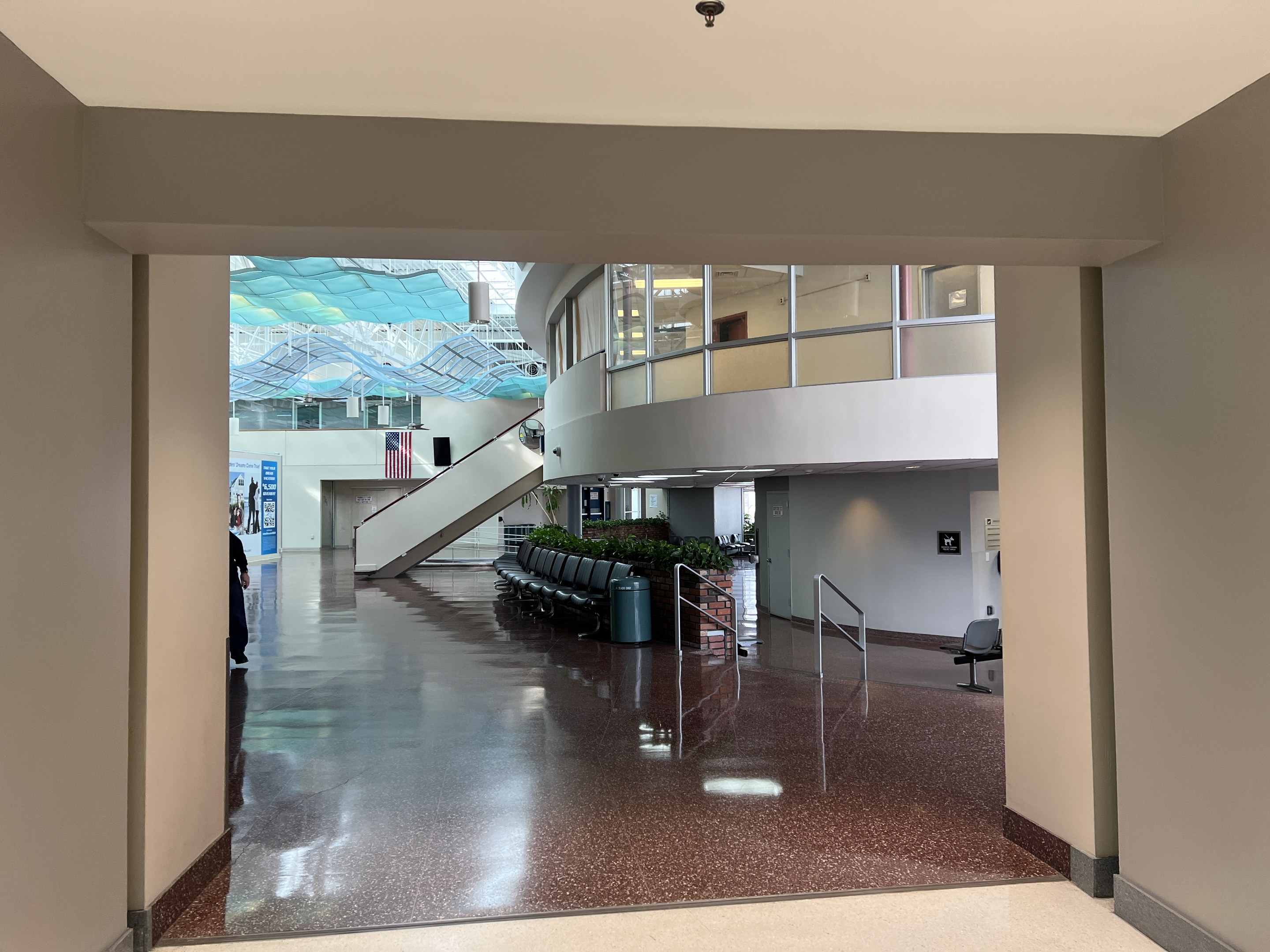Großer Eingangsbereich mit hohen Decken, zahlreichen Fenstern, einer zentralen Treppe mit Geländern, verstreuten Stühlen, Topfpflanzen, einem Mülleimer, einer Person, einem Schild und glänzendem Epoxidharz-Boden mit Deckenleuchten.