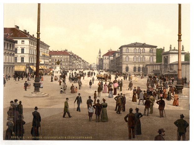 Ein altes Foto einer belebten Münchner Stadtstraße aus den frühen 1900er Jahren mit Passanten, einigen mit Schirmen, einem Pferdewagen im Vordergrund, Gebäuden, Bäumen und Pfählen im Hintergrund und Text am unteren Bildrand.