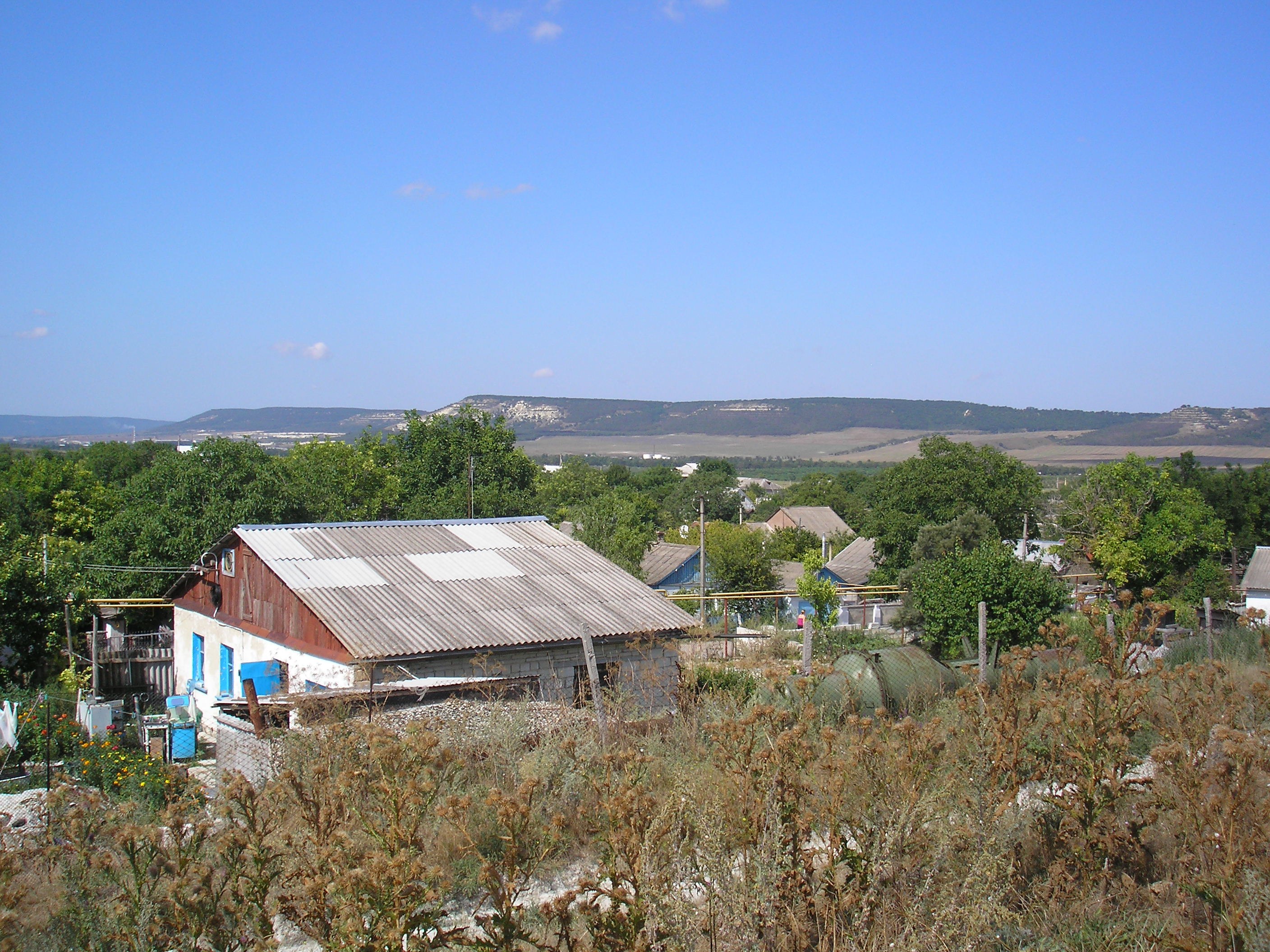 Ein kleines Dorf umgeben von Bäumen, Pflanzen und Strommasten, mit Hügeln und einem klaren blauen Himmel im Hintergrund.