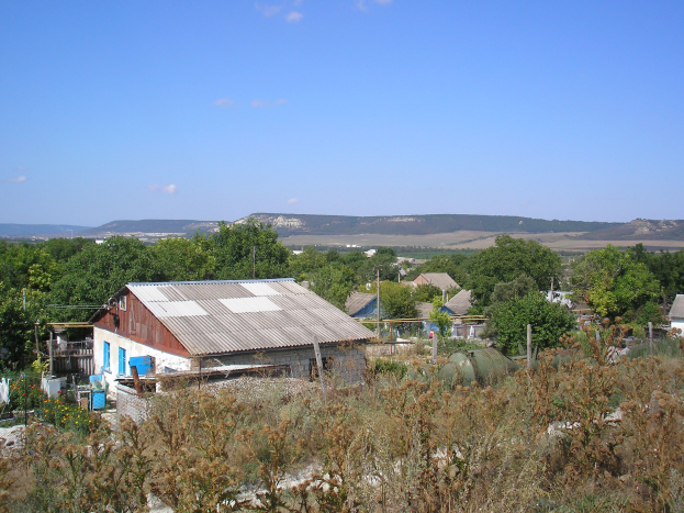 Ein kleines Dorf umgeben von Bäumen, Pflanzen und Strommasten, mit Hügeln und einem klaren blauen Himmel im Hintergrund.