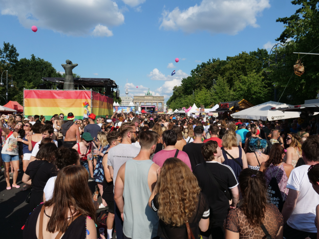 Eine große Menschenmenge marschiert auf einer Straße mit Zelten, Bäumen, Pfählen, Lichtern und einer Statue im Hintergrund, mit Gebäuden und einem Himmel mit Wolken und Ballons, während des Berliner Christopher Street Day.