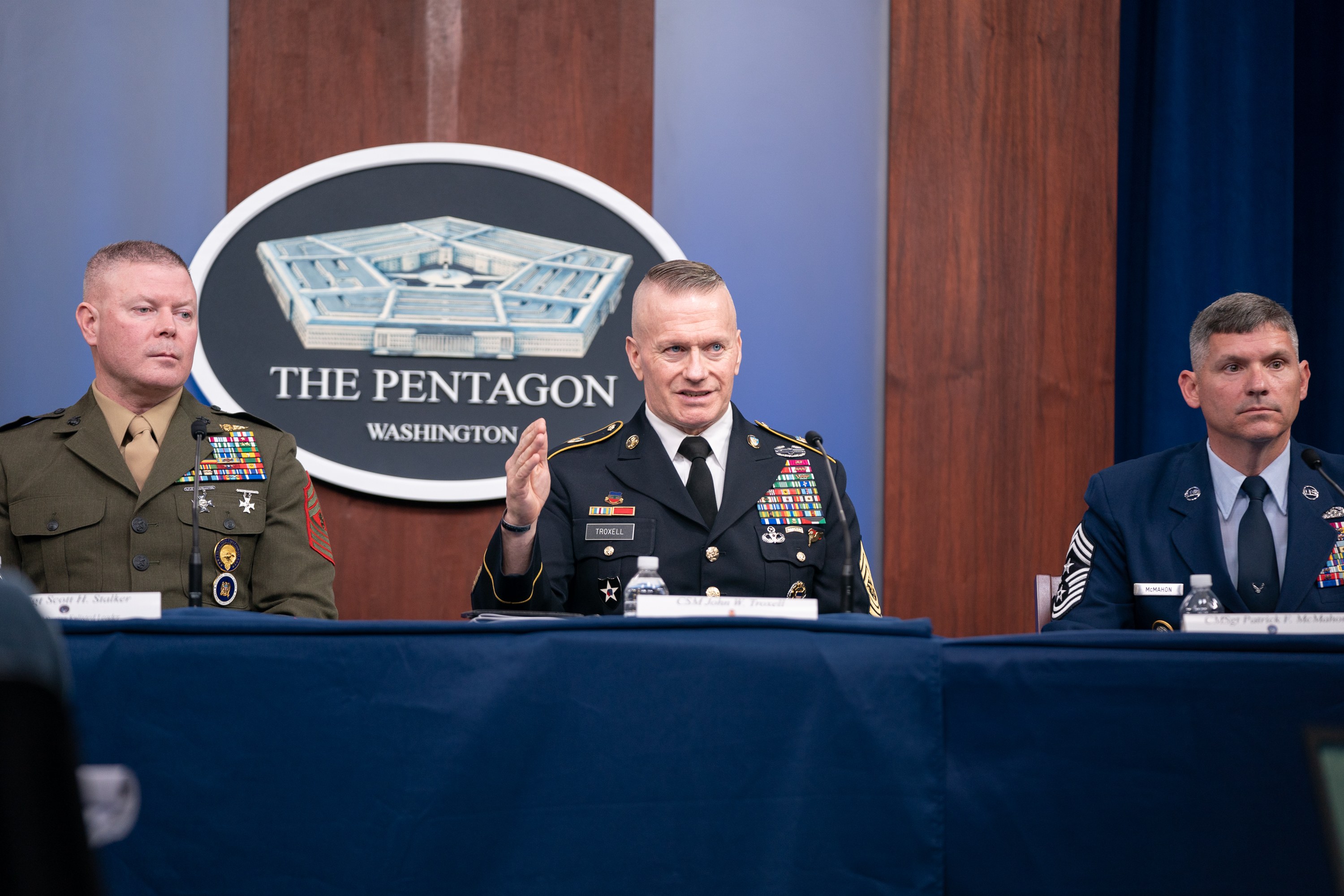 Drei Männer in militärischer Uniform sitzen an einem Tisch mit Namensschildern, Flaschen und Mikrofonen unter einer blauen Decke, mit einer Wand-Logo und einem Vorhang im Hintergrund.
