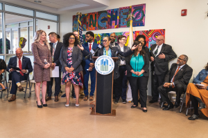 Gruppe von Menschen vor einem Rednerpult mit Mikrofon und einer Tafel mit der Aufschrift "Sancho-Stadtrat tritt dem Stadtrat bei", mit sitzenden und stehenden Teilnehmern, Flaggen, einem Feuerlöscher und Deckenleuchten.