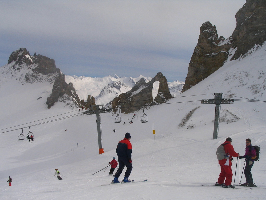Menschen in Pullovern sind auf dem Eis Ski fahren, mit einer Seilbahn, Bergen und einem bewölkten Himmel im Hintergrund.