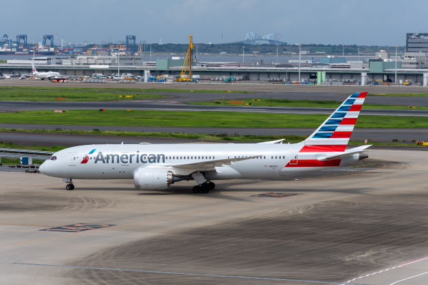 American Airlines Flugzeug auf einer Rollbahn mit Flughafeninfrastruktur, Gras, Fahrzeugen, Gebäuden, Polen, Kränen, Bäumen und einem klaren blauen Himmel.