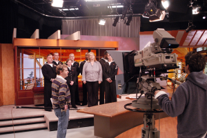 Gruppe von Menschen in einem Fernsehstudio mit einer Person, die eine Kamera hält, und Lampen an der Decke.