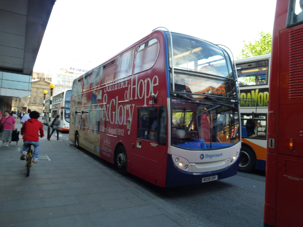 Eine Person f√§hrt mit einem Fahrrad auf einem Gehweg mit vielen Menschen, Bussen mit Text darauf, Geb√§uden und Laternen im Hintergrund.