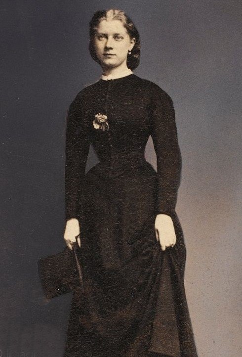 Ein Schwarz-Weiß-Foto einer älteren Frau in einem langen schwarzen Kleid mit einem hohen Kragen und langen Ärmeln, die eine Handtasche hält und deren Haar zu einem Dutt frisiert ist.