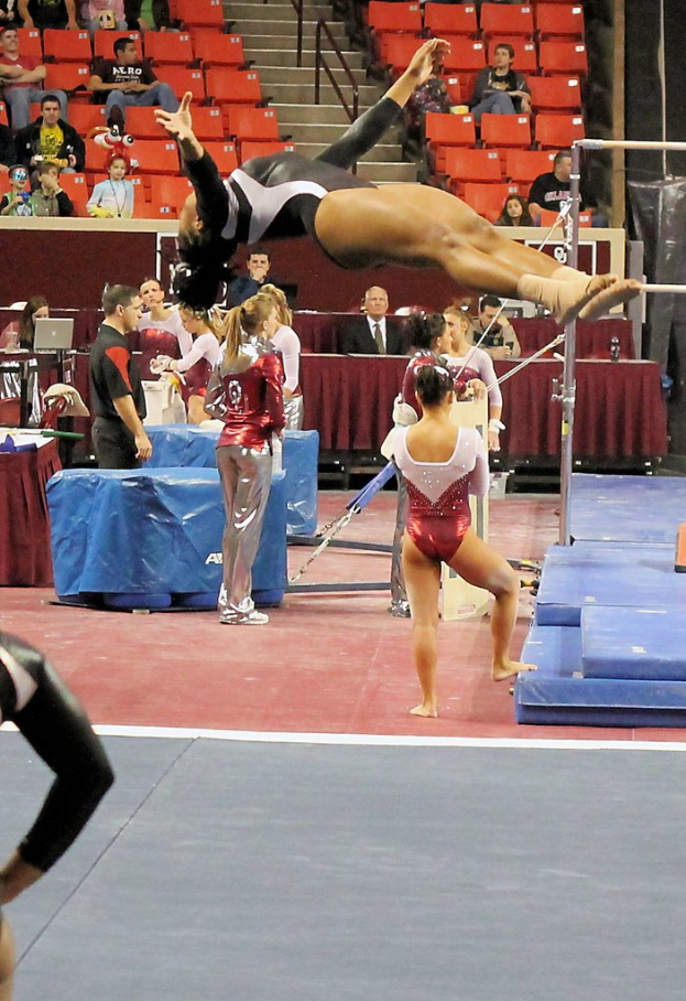 Personen sitzen und stehen in einer Turnhalle, wobei eine Person in der Mitte Gymnastik betreibt und Crashmatten auf der rechten Seite zu sehen sind.