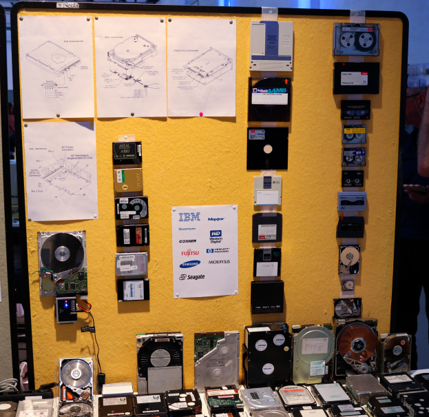 Ein Tisch voller verschiedener Festplatten, eine Person rechts stehend mit einem Gegenstand in der Hand und eine Tafel mit Papieren im Hintergrund.