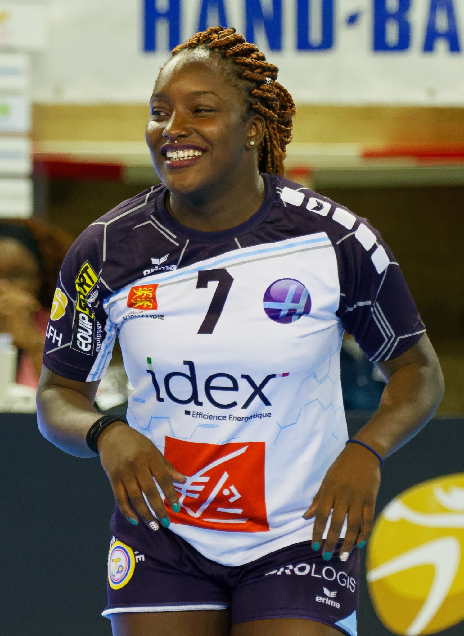 Eine professionelle Handballerin in einem lila und weißen Trikot steht auf einem Court, lächelt selbstbewusst, mit einem Hintergrund, der Menschen und eine Tafel mit der Aufschrift "FIVB FIVB Handball-Weltmeisterschaft 2019" zeigt.