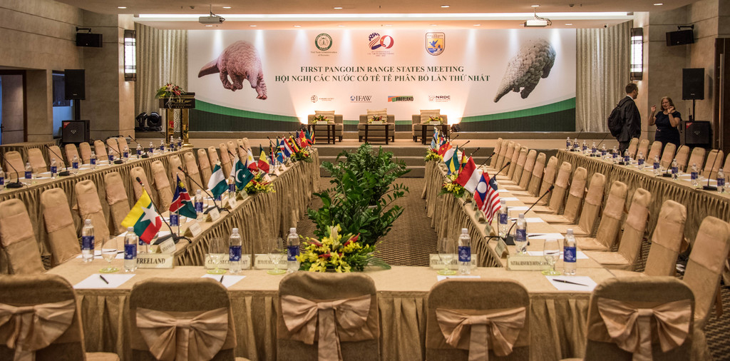 Eine B√ľhne mit Stühlen, Mikrofonen, Flaggen, Flaschen auf Tischen, einigen stehenden Menschen und einem Plakat.