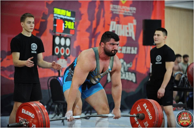 Ein Mann hebt eine Hantel bei einem Gewichtheben-Wettbewerb umgeben von Zuschauern, mit Wettkampfausrüstung und Hinweisschildern, die die 2018 World Weightlifting Championships anzeigen.