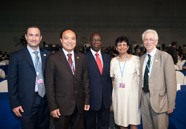Eine Gruppe von Männern in Anzügen und eine Frau in einem Kleid steht und lächelt im Vordergrund einer Halle, mit Menschen, die sitzen und stehen in der Mitte, Stühle, Tische und ein Mikrofon auf einem Tisch in der linken Ecke und eine Wand mit anderen Gegenständen im Hintergrund.