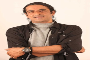 Ein Mann in einem schwarzen und aschgrauen Kleid mit überschlagenen Armen steht vor einem wei├čen Hintergrund, tr├Ągt eine Uhr und sieht nachdenklich aus.