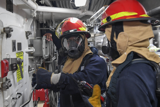 Zwei Feuerwehrleute in Schutzausrüstung, einschließlich Helmen und Gasmasken, arbeiten an einem Boot, mit einer Wand mit verschiedenen Gegenständen links und Rohren und anderen Gegenständen im Hintergrund.