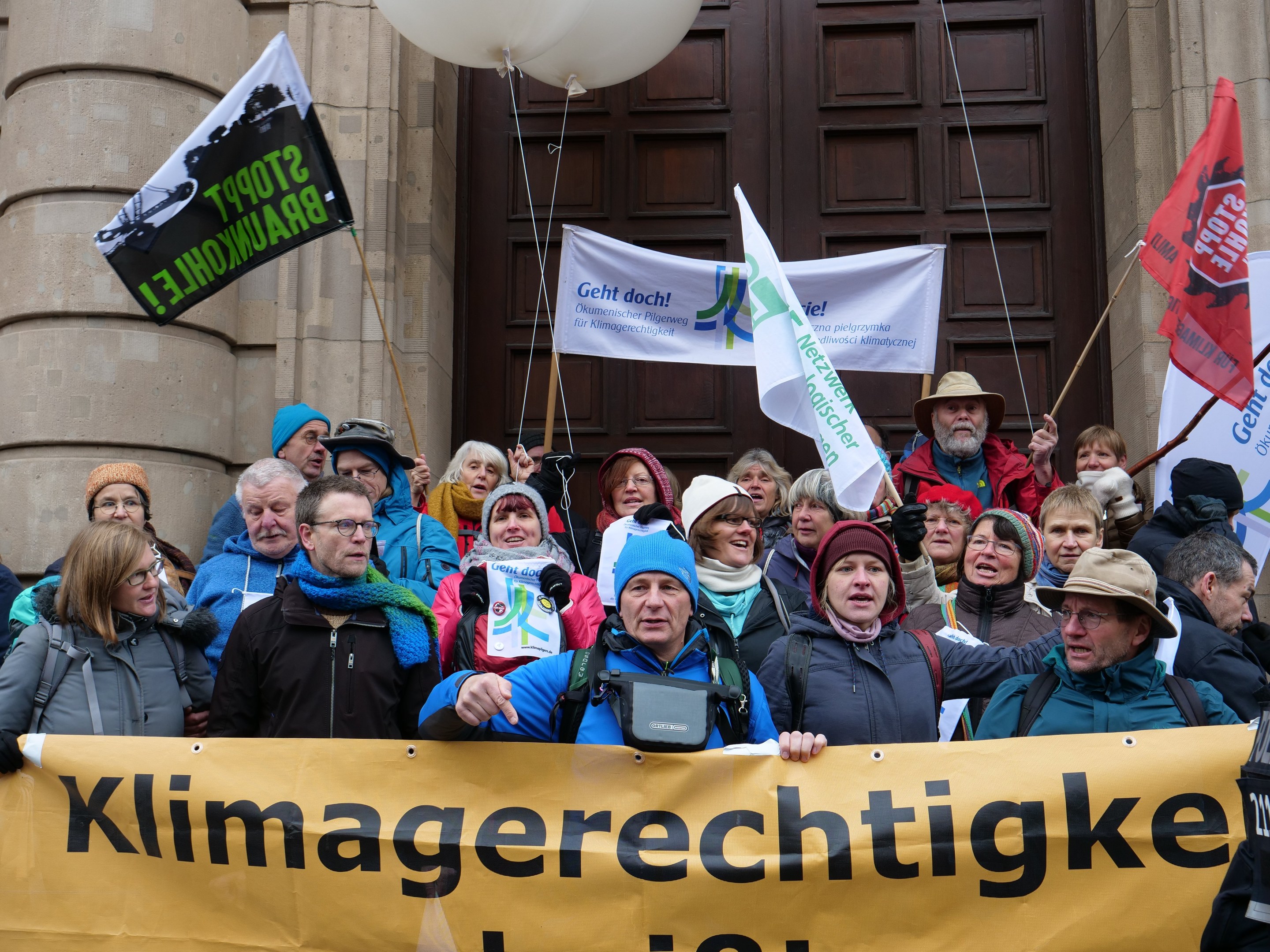 Gruppe von Menschen bei einer Klimademonstration vor einem Gebäude mit Säulen, die eine Fahne, Flaggen und Luftballons halten