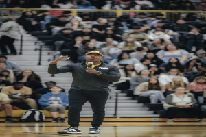 Ein Mann mit Mütze steht auf einem Basketballfeld und hält ein Mikrofon, hinter ihm sitzen Menschen auf einer Treppe und Taschen auf dem Boden.