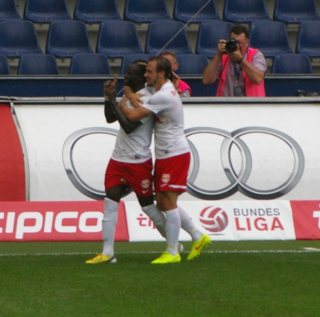 Zwei Fußballspieler feiern ein Tor auf einem Feld, umgeben von einer Gruppe von Menschen, mit einer Person, die eine Kamera hält, einem Banner, Stühlen und einer Wand im Hintergrund.