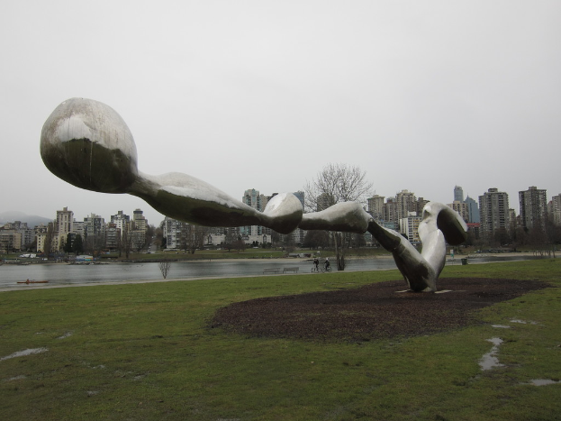Eine Skulptur im Vordergrund mit Erde und Gras, Gebäuden, Bäumen, Radfahrern und einer Wasserquelle in der Mitte, unter einem bewölkten Himmel.