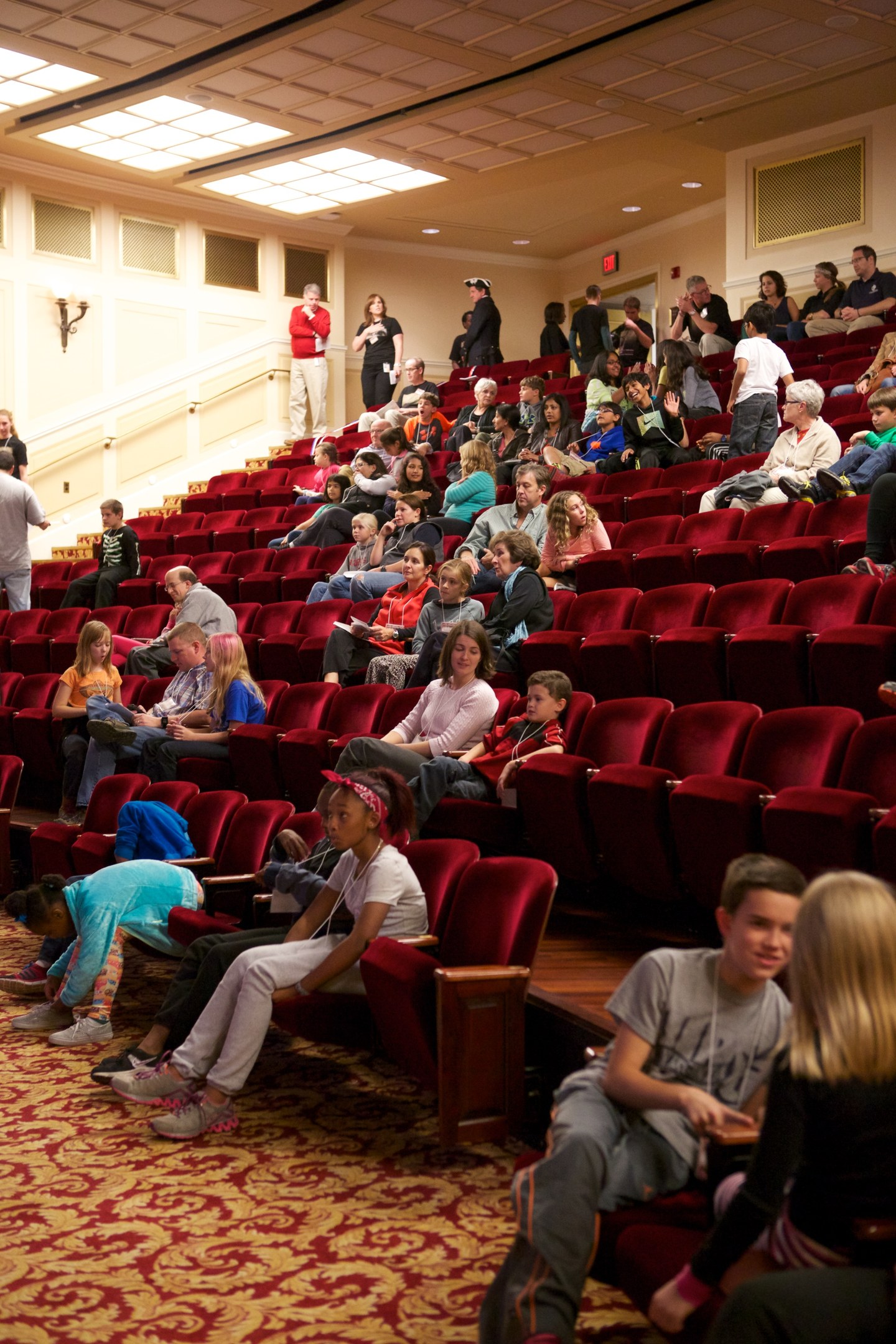 Eine große Gruppe von Menschen, darunter Kinder, die auf Stühlen sitzen und stehen, umgeben von verstreuten Stühlen, Wandlichtern und Deckenlichtern.