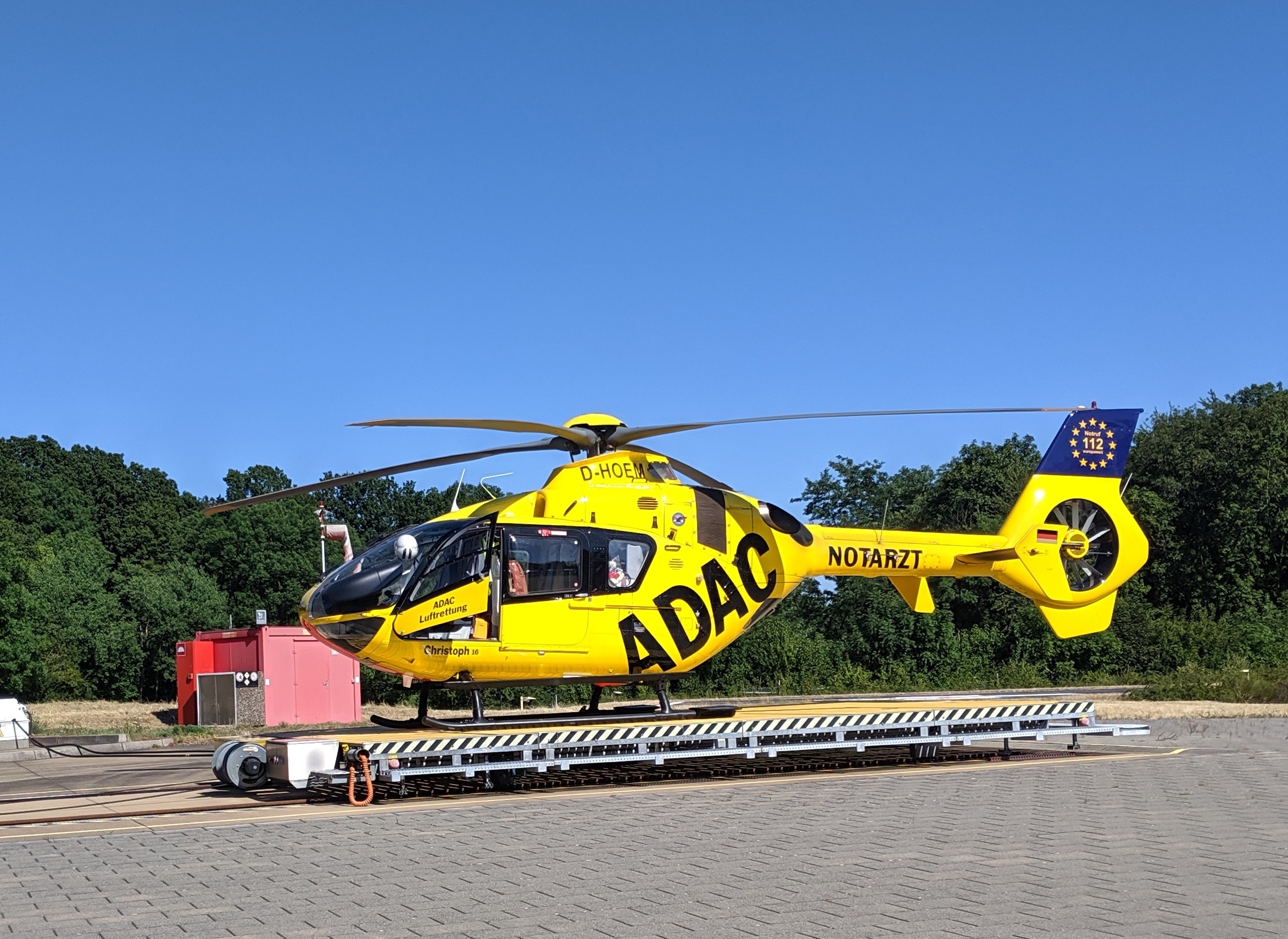 Ein gelber Hubschrauber steht auf dem Rollfeld eines Flughafens umgeben von Bäumen und einem klaren blauen Himmel, mit verschiedenen Gegenständen rund um den Hubschrauber verstreut.