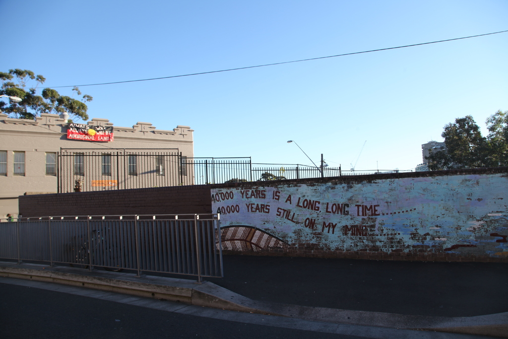 Eine Stadtansicht mit einer Straße, einem Geländer, Pflanzen, einer Wand im Vordergrund, Bäumen, Gebäuden, Eisenrahmen, einem Banner, einer Straßenlaterne und Kabel in der Mitte sowie Maschinen und Himmel im Hintergrund.