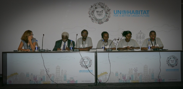 Sechs Personen in einem Konferenzsaal, eine Frau in einem grauen Kleid auf der linken Seite, eine Flasche auf dem rechten Tisch, ein weißes Banner im Hintergrund und zwei Tische mit Büchern, Mikrofonen und Flaschen im Vordergrund.
