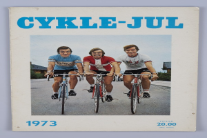 Drei Menschen auf Fahrrädern auf einer Straße umgeben von Häusern, Bäumen und einem klaren blauen Himmel, mit dem Text "Cykle-Jul 1973 - Cyclocross Magazine" oben und unten auf dem Bild.
