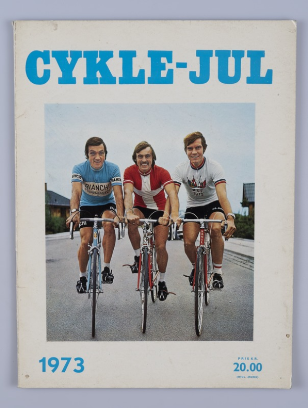Drei Menschen auf Fahrrädern auf einer Straße umgeben von Häusern, Bäumen und einem klaren blauen Himmel, mit dem Text "Cykle-Jul 1973 - Cyclocross Magazine" oben und unten auf dem Bild.