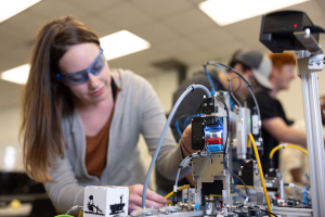 Eine Frau in Schutzbrille arbeitet an einem Roboter in einem Labor, umgeben von anderen, mit Werkzeugen und Komponenten auf dem Tisch unter Deckenbeleuchtung.