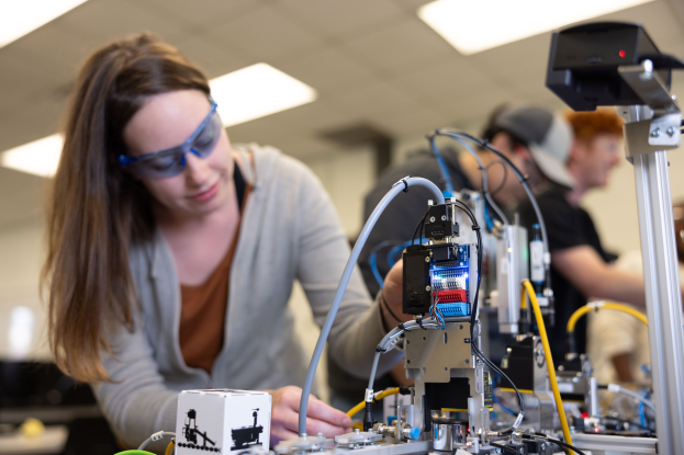 Eine Frau in Schutzbrille arbeitet an einem Roboter in einem Labor, umgeben von anderen, mit Werkzeugen und Komponenten auf dem Tisch unter Deckenbeleuchtung.