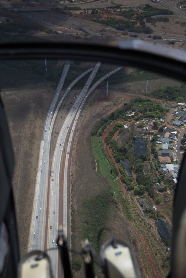 Ein Blick aus dem Flugzeugfenster, der Straßen mit Fahrzeugen, Bäumen, Masten und Gebäuden darunter zeigt.