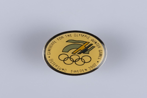 Ein schwarzes Abzeichen mit goldenem Olympic Winter Games-Logo und weißer Schrift auf einer weißen Oberfläche.