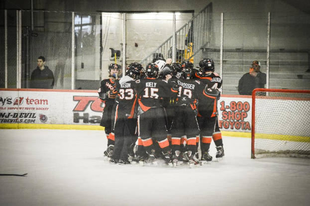 Eine Gruppe von Menschen in Schlittschuhen, Handschuhen und Helmen steht auf einem Eisfläche mit Absperrungen und Glastüren dahinter, ein Hockey-Tor mit einem Netz auf der rechten Seite und einen Hockey-Schläger auf der linken Seite.