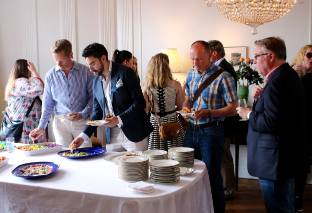 Eine Gruppe von Menschen sitzt um einen weißen Tisch mit Essen und Tellern, mit dekorativen Lampen und einer Deckenleuchte im Hintergrund.