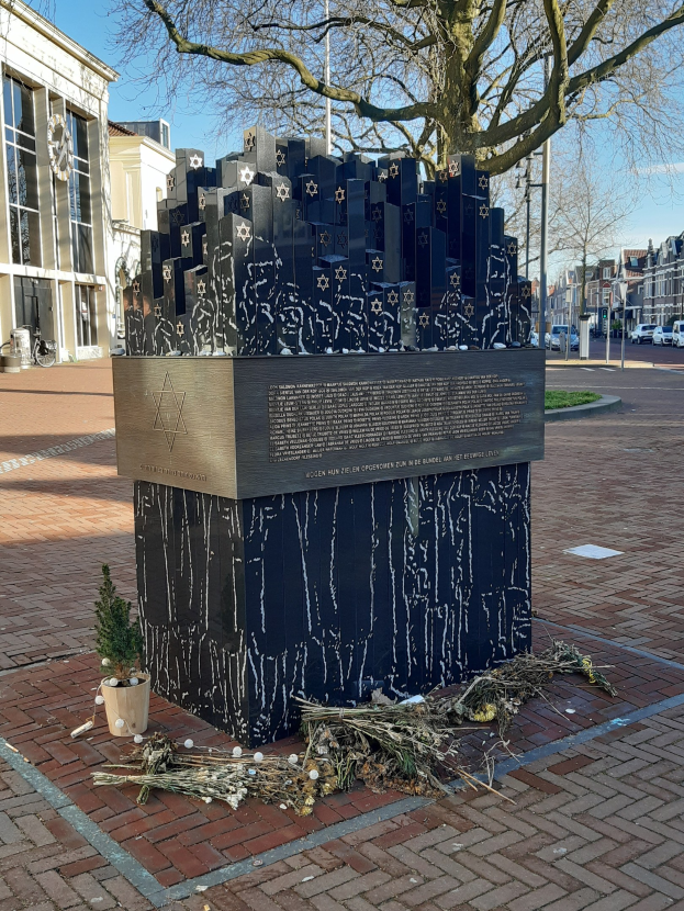 Denkmal in einem Stadtplatz für Holocaust-Opfer mit einem Baum, Topfpflanze, verstreuten trockenen Blättern, umgeben von Gebäuden, Fahrzeugen, Versorgungsmästen und Bäumen unter dem Himmel.